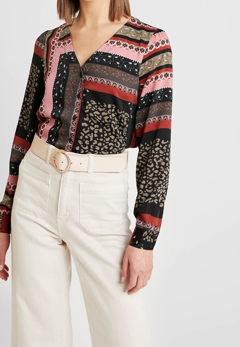 Femme portant un chemisier multicolore à motifs floraux et animaux, rentré dans un pantalon blanc taille haute avec une large ceinture.