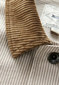 Tissu beige et marron rayé avec col en velours côtelé marron, bouton noir et étiquette blanche partielle montrant un texte et un logo.