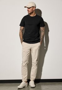 Camiseta negra de manga corta, pantalones ajustados beige y zapatillas de color claro. El diseño incluye tatuajes en los brazos y una gorra beige.