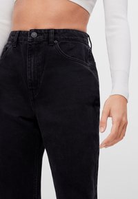 Jeans de mezclilla negra de tiro alto con una textura suave, que cuentan con un cierre de botón, lazos para el cinturón y bolsillos laterales.