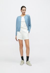 Cardigan tricoté bleu clair porté sur un haut blanc et un short blanc. Porté avec des chaussettes noires et des baskets beige. Texture lisse, design simple.