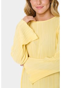 Yellow pleated dress featuring a round neckline and loose long sleeves, with a textured fabric creating a flowing, layered effect.
