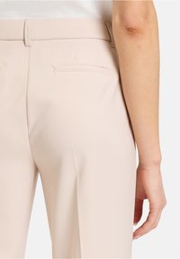 Světlé béžové kalhoty na míru s hladkým materiálem. Design obsahuje plochý přední díl, dvě zadní kapsy a poutka na opasek. Ruka odpočívající vedle kalhot.
