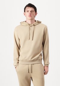 Hanorac bej cu șnur, manșete și tiv cu structuri în relief. Prezentă un logo pe piept. Asortat cu pantaloni jogger bej. Textură netedă a materialului.