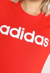 T-shirt in cotone rosso con scollatura a girocollo, caratterizzato da una scritta "adidas" in bianco audace sul davanti. Texture liscia, design aderente.