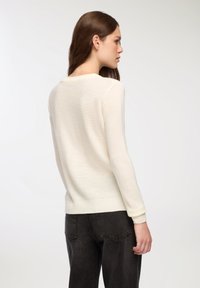 Jasna kremowa sweter z długimi rękawami, wykonany z miękkiego dzianinowego materiału, z okrągłym dekoltem i ściągaczem, zestawiony z ciemnoszarymi prostymi dżinsami.