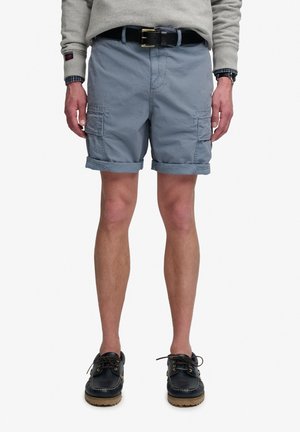Mann trägt hochgekrempelte blaue Cargoshorts, schwarzen Gürtel, grauen Pullover und schwarze Bootsschuhe, steht vor weißem Hintergrund.