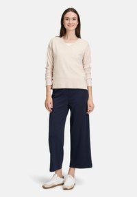 Beiger V-Ausschnitt-Pullover, lockere Passform, kombiniert mit marineblauen weiten Hosen und weißen Sneakers. Natürliche Stoffstruktur, klare Linien und schlichtes Design.