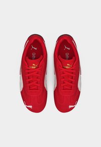 Ζευγάρι κόκκινα αθλητικά παπούτσια Puma με λευκό λογότυπο και χρυσή εμπορική σήμανση, σε θέαση από πάνω σε λευκό φόντο.