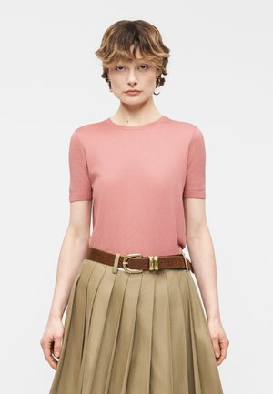 Ženska s kratkimi valovitimi lasmi nosi roza top z kratkimi rokavi, rjav pas in bež plisirano krilo na enostavni beli podlagi.