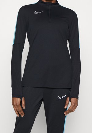 Czarna sportowa bluzka Nike z długim rękawem, półzamkiem i jasnoniebieskimi paskami na rękawach, noszona z dopasowanymi czarnymi spodniami z niebieskimi paskami po bokach.