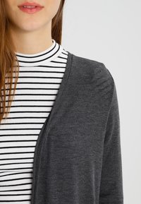 Cárdigan gris carbón sobre un suéter de cuello alto a rayas blancas y negras. El cárdigan presenta un escote en V y una textura de tela suave.