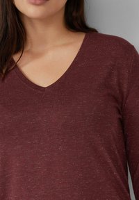 Burgunderfarbene V-Ausschnitt langärmlige Bluse aus strukturiertem Strickstoff mit dezenten Einsprengseln. Weiches Material, figurbetonte Silhouette und schlichtes Design.