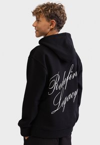 Juodas gobtuvas, pagamintas iš minkštos medžiagos, su dideliu baltu užrašų tekstu ant nugaros, sakant „Redefin Legacy“. Standartinio kirpimo, turintis kišenę kanguru.