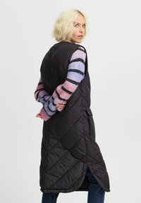 Svart, vadderad lång väst med hög krage och diagonala sömmar; bärs över en flerfärgad randig tröja med pastellnyanser.