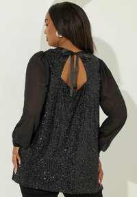 Svart paljettopp med genomskinliga puffärmar, nyckelhålsutskärning på ryggen och banddetalj. Mjuk textur med en glittrande yta.