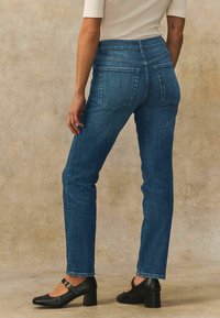 Donkerblauwe denim jeans met een getailleerde pasvorm, vijfzakkenontwerp en subtiele stikdetails. Gecombineerd met zwarte schoenen met blokhak.