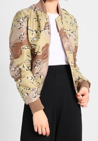 Kamouflage bomberjacka i beige och brunt med svarta detaljer, dragkedjestängning, ribbade manschetter och en slät yta. Buren �över en vit topp.