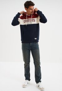 Sudadera de bloques de color en azul marino y burdeos con el texto rojo "CHEEKS ORIGINAL desde 1974", que incluye un bolsillo frontal y capucha con cordones. Se lleva con vaqueros.