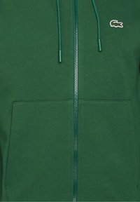 Sweat à capuche vert zippé avec poches avant, cordons et petit logo crocodile brodé sur la poitrine.