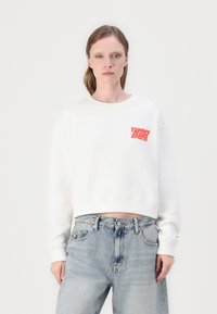 Bílá zkrácená mikina s kulatým výstřihem a žebrovanými manžetami. Na levé hrudi má červené logo "TOMMY JEANS". Nosí se s lehkou modrou džínovinou.