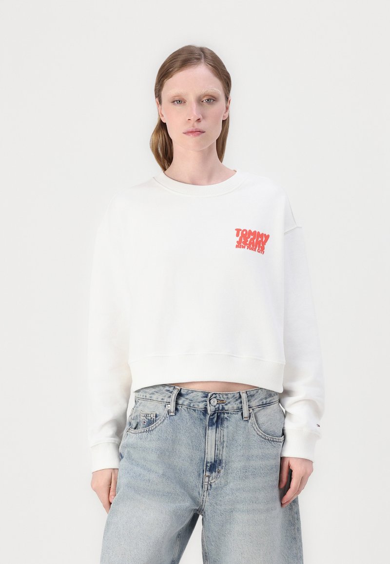 Valge lühendatud sweatshirt ümmarguse kaelusega ja ribidega varrukatega. Vasakul rinnal on punane "TOMMY JEANS" logo. Kanna heledate siniste teksadega.