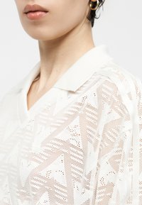 Κοντινό πλάνο ενός ατόμου που φοράει λευκό πουκάμισο από δαντέλα με σχέδια και γιακά, καθώς και χρυσούς κρίκους στα αυτιά, δείχνοντας τα χείλη και τον λαιμό.