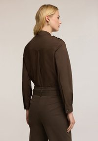 Hnědá košile s límečkem a dlouhými rukávy, se středovým švem na zádech a knoflíky na ramenou; v kombinaci s odpovídajícími hnědými kalhotami.