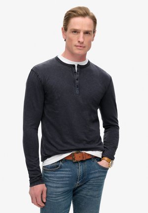 Camisa henley de manga larga en azul marino oscuro, con una tapeta de tres botones, tejido texturizado y escote redondeado, llevada sobre una camiseta blanca y jeans azules.