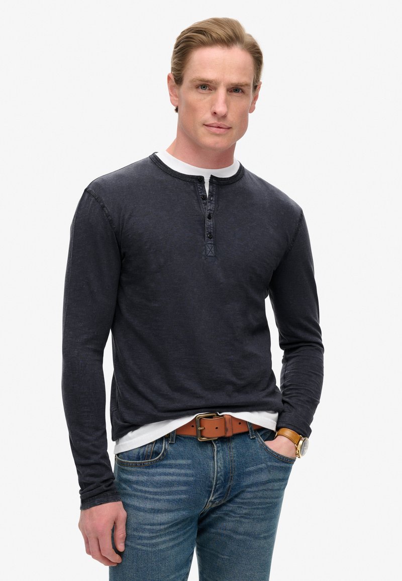 Dunkelblaues Langarm-Henley-Shirt mit einer Dreiknopf-Leiste, strukturiertem Stoff und rundem Ausschnitt, getragen über einem weißen T-Shirt und blauen Jeans.