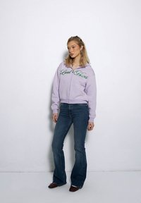 Fioletowa bluza z kapturem na zamek z haftem "Local Heroes" w kolorze zielonym, w połączeniu z ciemnoniebieskimi spodniami dzwonami i brązowymi butami. Gładka struktura materiału.