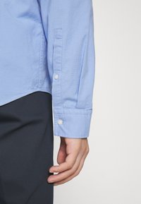 Camisa de manga larga azul claro con puños de botón. Tela de algodón, textura suave y un dobladillo curvado visible en el lateral de la camisa.
