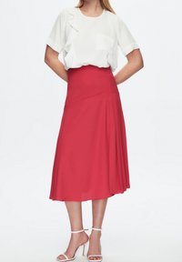 Falda roja en línea A con pliegues, combinada con una blusa blanca con un detalle de volantes y un bolsillo. Llevada con tacones blancos con tiras.