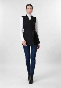 Γυναίκα όρθια, φοράει μαύρο αμάνικο παλτό με ζώνη πάνω από λευκό ζιβάγκο, σκούρο μπλε στενά τζιν και μαύρα μποτάκια αστραγάλου σε απλό φόντο.