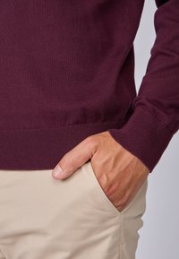Pull en maille bourgogne avec des poignets et un ourlet côtelés, porté sur un pantalon beige, avec une main reposant dans la poche du pantalon.