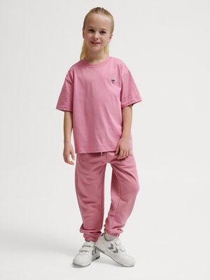 Młoda dziewczyna stoi, ubrana w luźny różowy t-shirt, pasujące różowe spodnie dresowe oraz białe tenisówki na jednolitym białym tle.