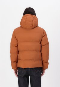 Hnědá péřová bunda s velkou kapucí, prošívaným designem a elastickými manžetami, doplněná tmavými džínami z denimu. Jednoduché, čisté linie.