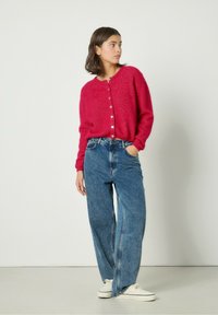 Cárdigan de punto fucsia con cuello redondo y botones, combinado con unos vaqueros azules sueltos y zapatillas blancas. Textura suave, corte casual.