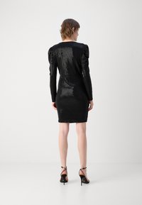 Robe noire à sequins avec manches longues et épaules bouffantes, silhouette ajustée et ourlet au genou. Associée à des escarpins noirs à lanières.