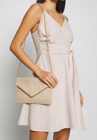 Borsa clutch beige con superficie texturizzata e tracolla a catena dorata, tenuta contro un vestito beige chiaro con un design avvolgente e lusinghiero.
