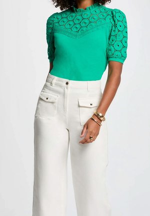 Femme portant un chemisier vert en dentelle à manches courtes et un pantalon blanc taille haute avec des poches à rabat à l'avant, accessoirisée avec des bracelets et une bague en or.