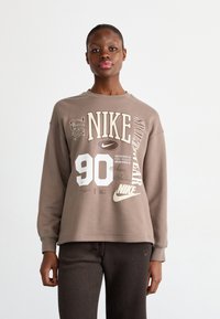 Brūns Nike sporta krekls ar grafisku dizainu, kurā ir krēmkrāsas teksts, tostarp "90" un "sportwear". Garas piedurknes ar ribota tipa aprocēm. Mīksta tekstūra.