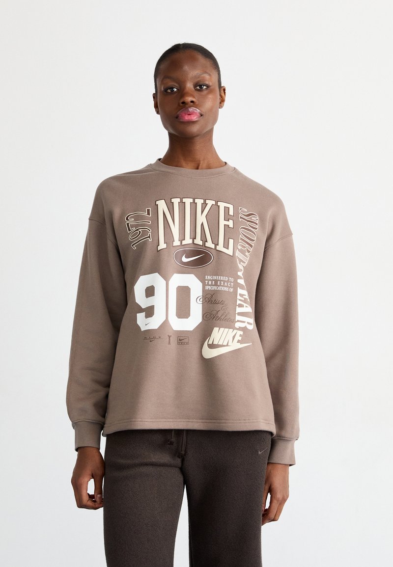 Brūns Nike sporta krekls ar grafisku dizainu, kurā ir krēmkrāsas teksts, tostarp "90" un "sportwear". Garas piedurknes ar ribota tipa aprocēm. Mīksta tekstūra.