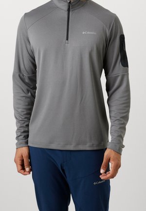 Mann trägt einen grauen Langarm-Halbzip-Pullover mit schwarzer Arm-Tasche und marineblauen Hose, beide mit Columbia-Logos.