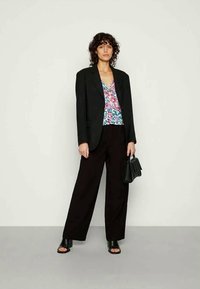 Mulher com cabelo encaracolado, vestindo um blazer preto, uma blusa floral, calças largas pretas e sapatilhas abertas pretas, segurando uma bolsa preta, de pé contra uma parede lisa.