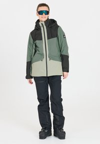 Skijack in donker groen en zwart, met een ritssluiting aan de voorkant, een capuchon en grote zakken. Gecombineerd met zwarte skibroek en skischoenen.