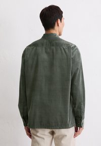 Camicia in corduroy verde, maniche lunghe, polsini con bottoni, colletto, superficie testurizzata, vestibilità comoda, esposta da dietro con pantaloni beige.