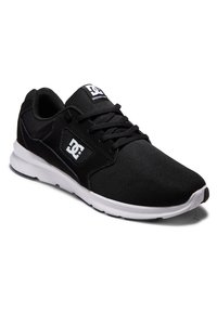 Chaussure de sport noire avec tige en mesh et superpositions synthétiques. Présente une semelle blanche, des lacets, et un logo sur la languette et le côté. Texture lisse.