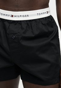 Άνδρας κορμός που φοράει μαύρα μποξεράκια Tommy Hilfiger με λευκή ελαστική μέση με το όνομα και το λογότυπο της μάρκας.