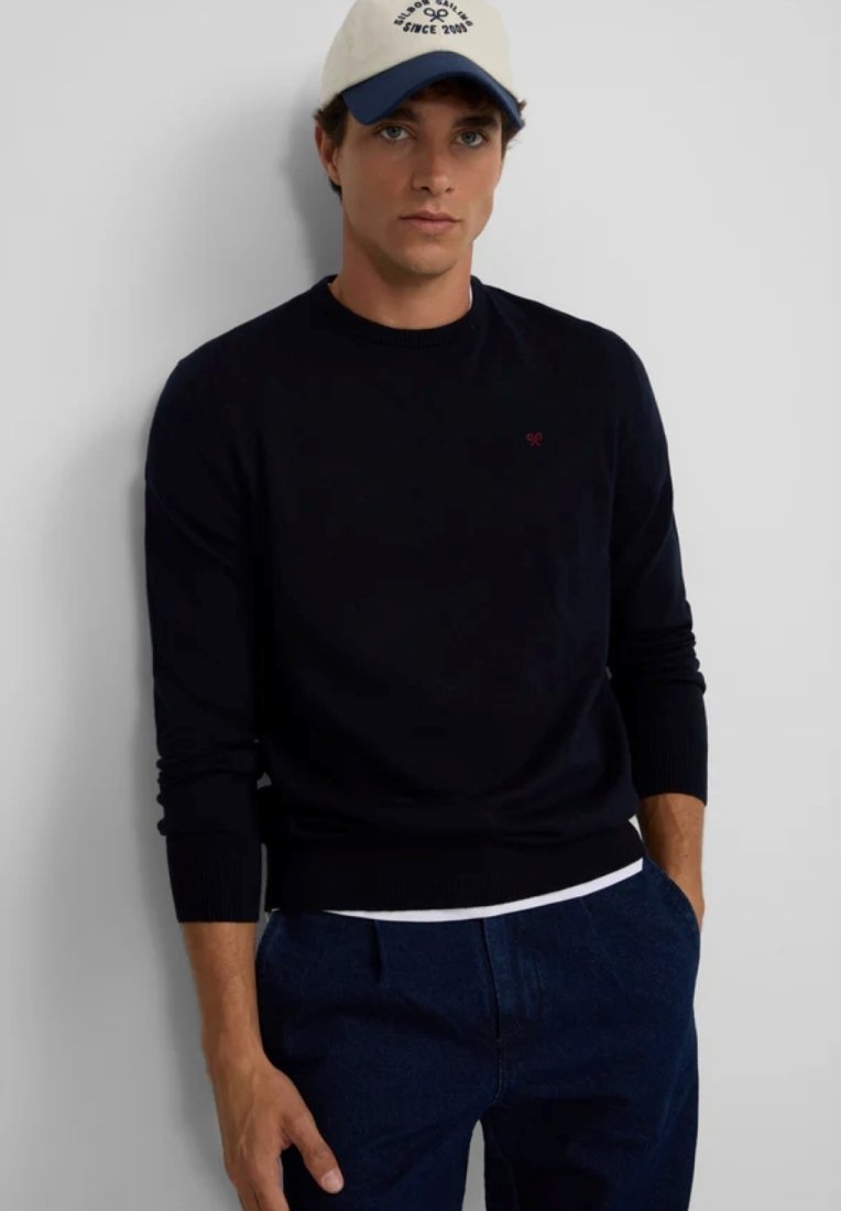 Jersey de punto navy con mangas largas, puños acanalados y cuello redondo. Presenta un pequeño logotipo rojo en el pecho y un dobladillo de camisa blanca superpuesto.